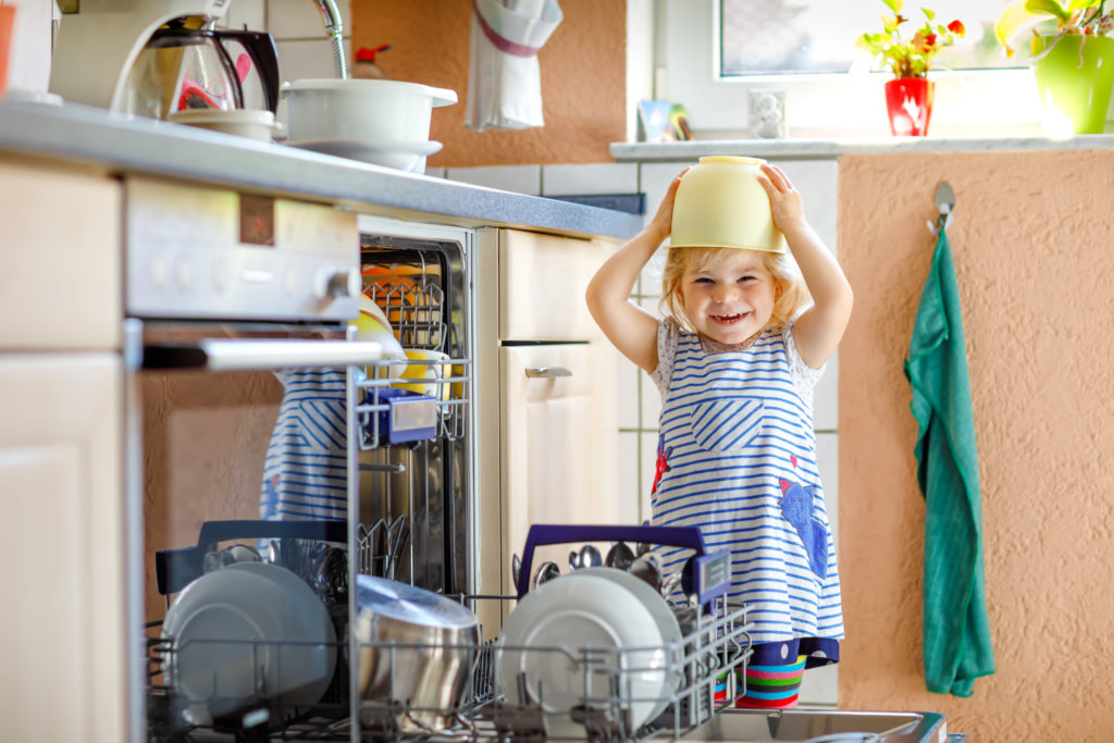 Little adorable child helping to unload dishwasher. Funny happy toddler girl standing in the kitchen, holding dishes and putting a bowl on head. Healthy kid at home.